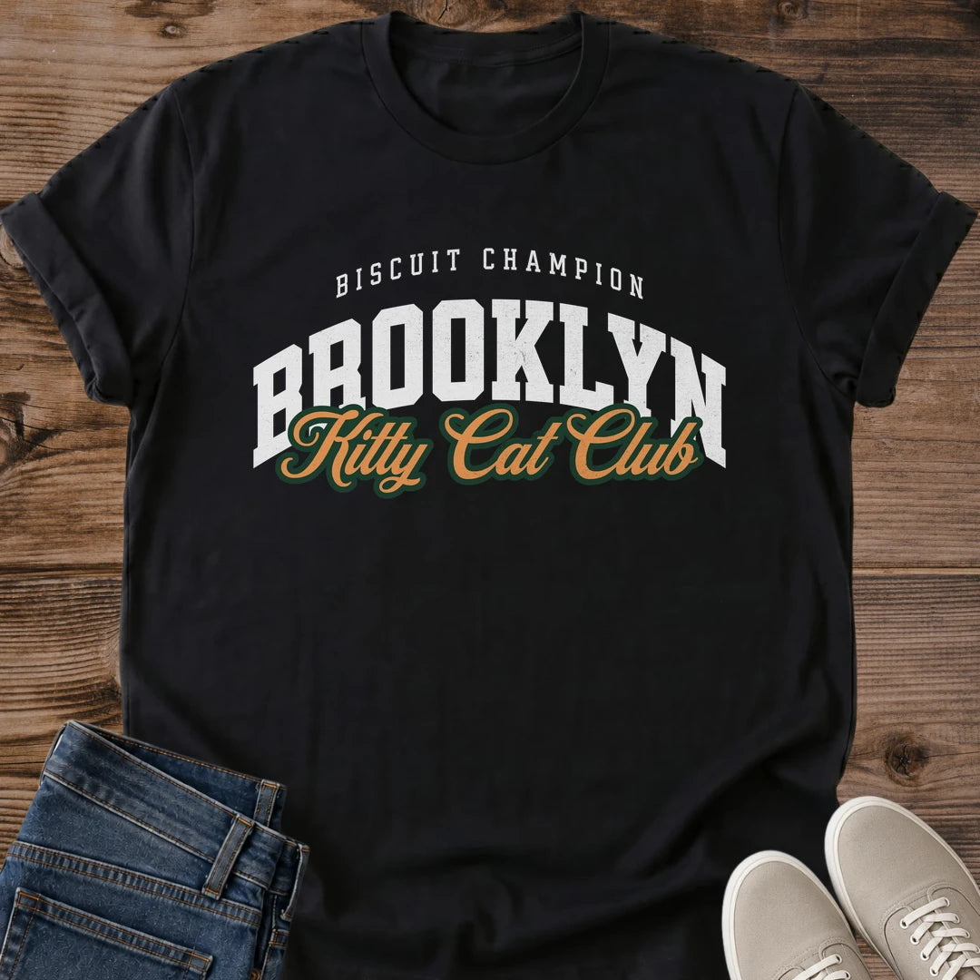 Black Brooklyn Kitty Cat Club T-Shirt, a Gildan 64000 Softstyle unisex crewneck with rolled short sleeves. The chest print pairs small arched white "BISCUIT CHAMPION" text with large white "BROOKLYN" varsity letters and overlapping gold "Kitty Cat Club" script outlined in dark green. Front flat lay on rustic brown wood planks, with folded dark denim jeans at lower left and light gray lace-up sneakers at lower right.