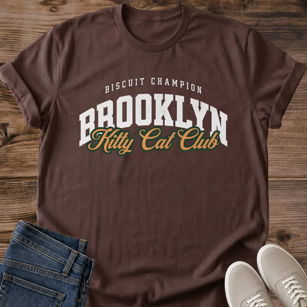 Dark Chocolate deep brown Brooklyn Kitty Cat Club T-Shirt, a Gildan 64000 Softstyle unisex crewneck with rolled short sleeves. The chest print pairs small arched white "BISCUIT CHAMPION" text with large white "BROOKLYN" varsity letters and overlapping gold "Kitty Cat Club" script outlined in dark green. Front flat lay on rustic brown wood planks, with folded dark denim jeans at lower left and light gray lace-up sneakers at lower right.