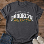 Dark Heather charcoal gray Brooklyn Kitty Cat Club T-Shirt, a Gildan 64000 Softstyle unisex crewneck with rolled short sleeves. The chest print pairs small arched white "BISCUIT CHAMPION" text with large white "BROOKLYN" varsity letters and overlapping gold "Kitty Cat Club" script outlined in dark green. Front flat lay on rustic brown wood planks, with folded dark denim jeans at lower left and light gray lace-up sneakers at lower right.