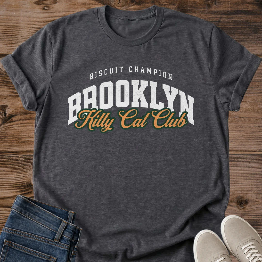 Dark Heather charcoal gray Brooklyn Kitty Cat Club T-Shirt, a Gildan 64000 Softstyle unisex crewneck with rolled short sleeves. The chest print pairs small arched white "BISCUIT CHAMPION" text with large white "BROOKLYN" varsity letters and overlapping gold "Kitty Cat Club" script outlined in dark green. Front flat lay on rustic brown wood planks, with folded dark denim jeans at lower left and light gray lace-up sneakers at lower right.