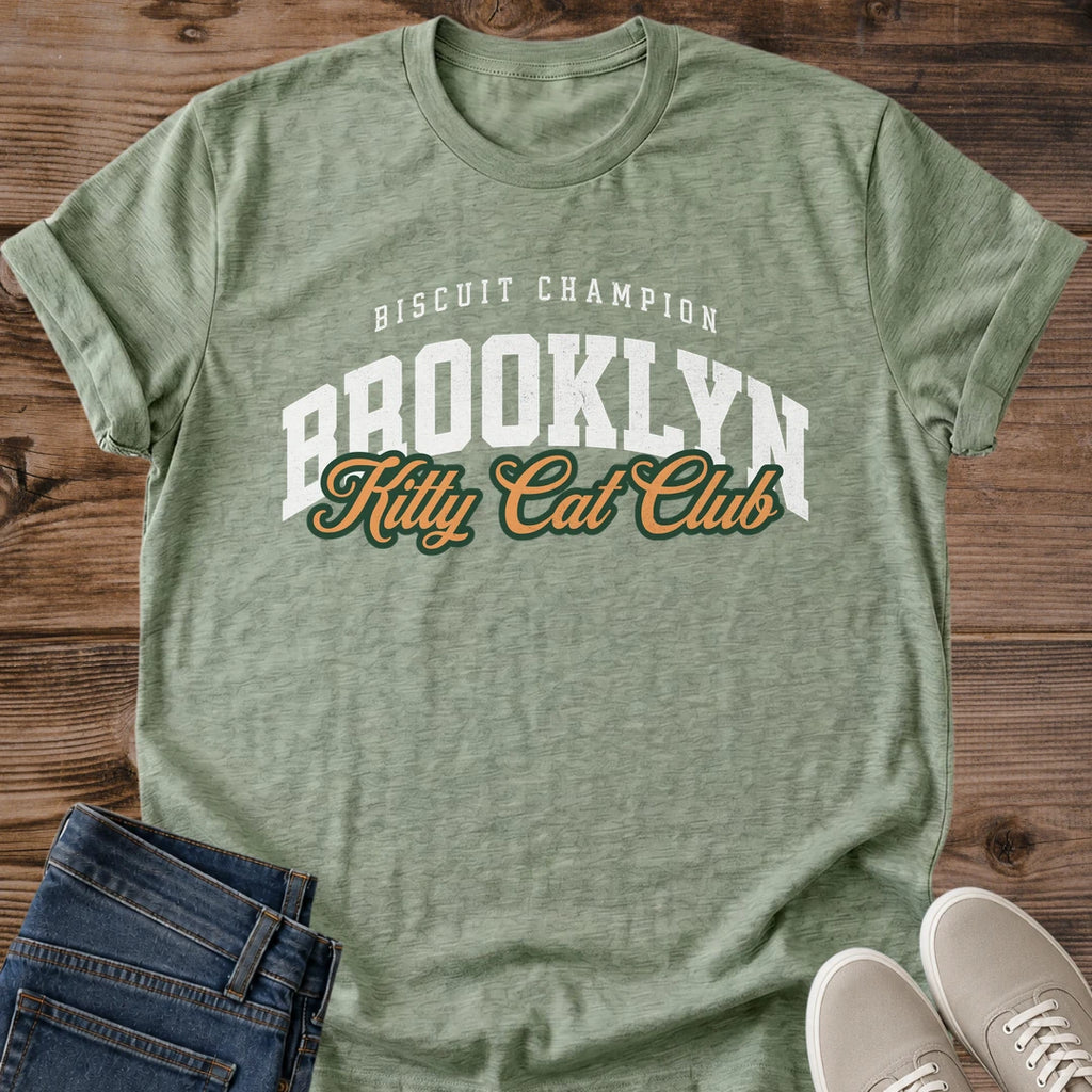 Heather Military Green with a mottled green texture Brooklyn Kitty Cat Club T-Shirt, a Gildan 64000 Softstyle unisex crewneck with rolled short sleeves. The chest print pairs small arched white "BISCUIT CHAMPION" text with large white "BROOKLYN" varsity letters and overlapping gold "Kitty Cat Club" script outlined in dark green. Front flat lay on rustic brown wood planks, with folded dark denim jeans at lower left and light gray lace-up sneakers at lower right.