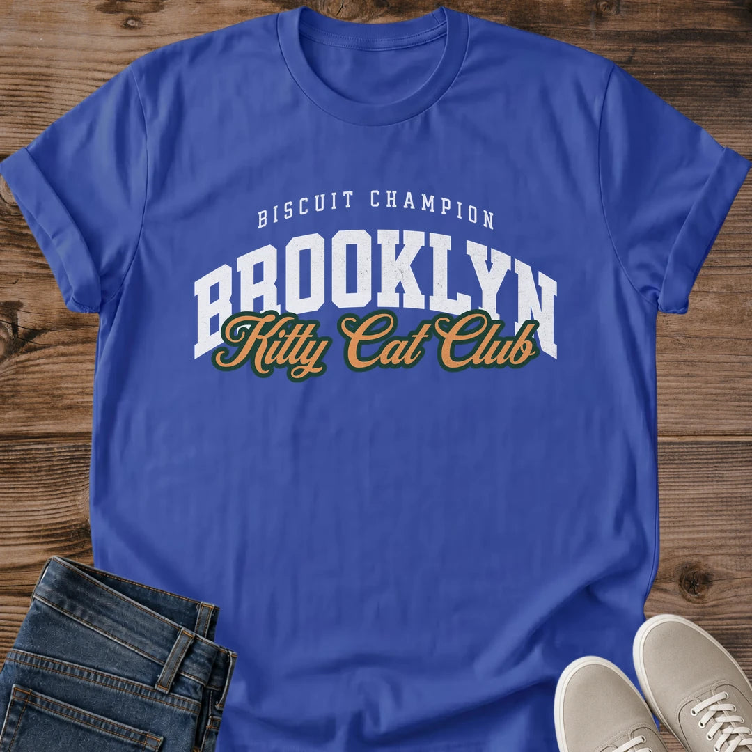 Metro Blue vivid blue Brooklyn Kitty Cat Club T-Shirt, a Gildan 64000 Softstyle unisex crewneck with rolled short sleeves. The chest print pairs small arched white "BISCUIT CHAMPION" text with large white "BROOKLYN" varsity letters and overlapping gold "Kitty Cat Club" script outlined in dark green. Front flat lay on rustic brown wood planks, with folded dark denim jeans at lower left and light gray lace-up sneakers at lower right.