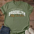 Military Green olive Brooklyn Kitty Cat Club T-Shirt, a Gildan 64000 Softstyle unisex crewneck with rolled short sleeves. The chest print pairs small arched white "BISCUIT CHAMPION" text with large white "BROOKLYN" varsity letters and overlapping gold "Kitty Cat Club" script outlined in dark green. Front flat lay on rustic brown wood planks, with folded dark denim jeans at lower left and light gray lace-up sneakers at lower right.
