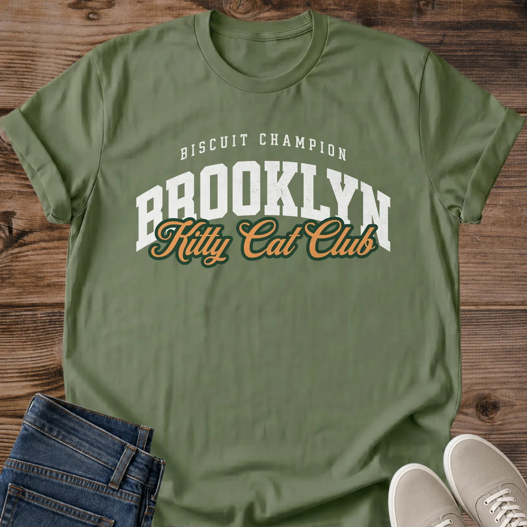 Military Green olive Brooklyn Kitty Cat Club T-Shirt, a Gildan 64000 Softstyle unisex crewneck with rolled short sleeves. The chest print pairs small arched white "BISCUIT CHAMPION" text with large white "BROOKLYN" varsity letters and overlapping gold "Kitty Cat Club" script outlined in dark green. Front flat lay on rustic brown wood planks, with folded dark denim jeans at lower left and light gray lace-up sneakers at lower right.