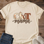 Sand Cat Mama graphic T-shirt on a wood surface, featuring a seated leopard-spotted cat, floral C, leopard-print T and brown mama script.