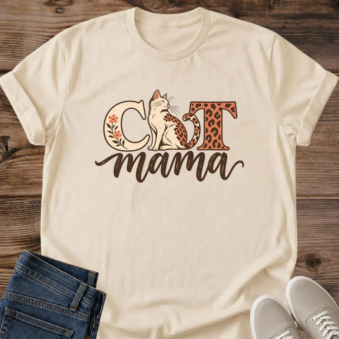 Sand Cat Mama graphic T-shirt on a wood surface, featuring a seated leopard-spotted cat, floral C, leopard-print T and brown mama script.