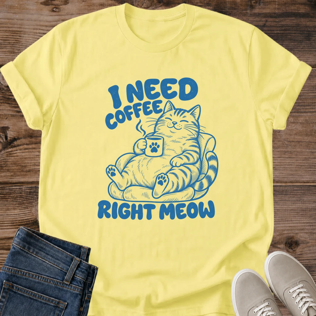 Cornsilk yellow T-shirt flat lay with blue lettering reading I Need Coffee Right Meow and a tabby lounging in an armchair holding a steaming paw-print mug.