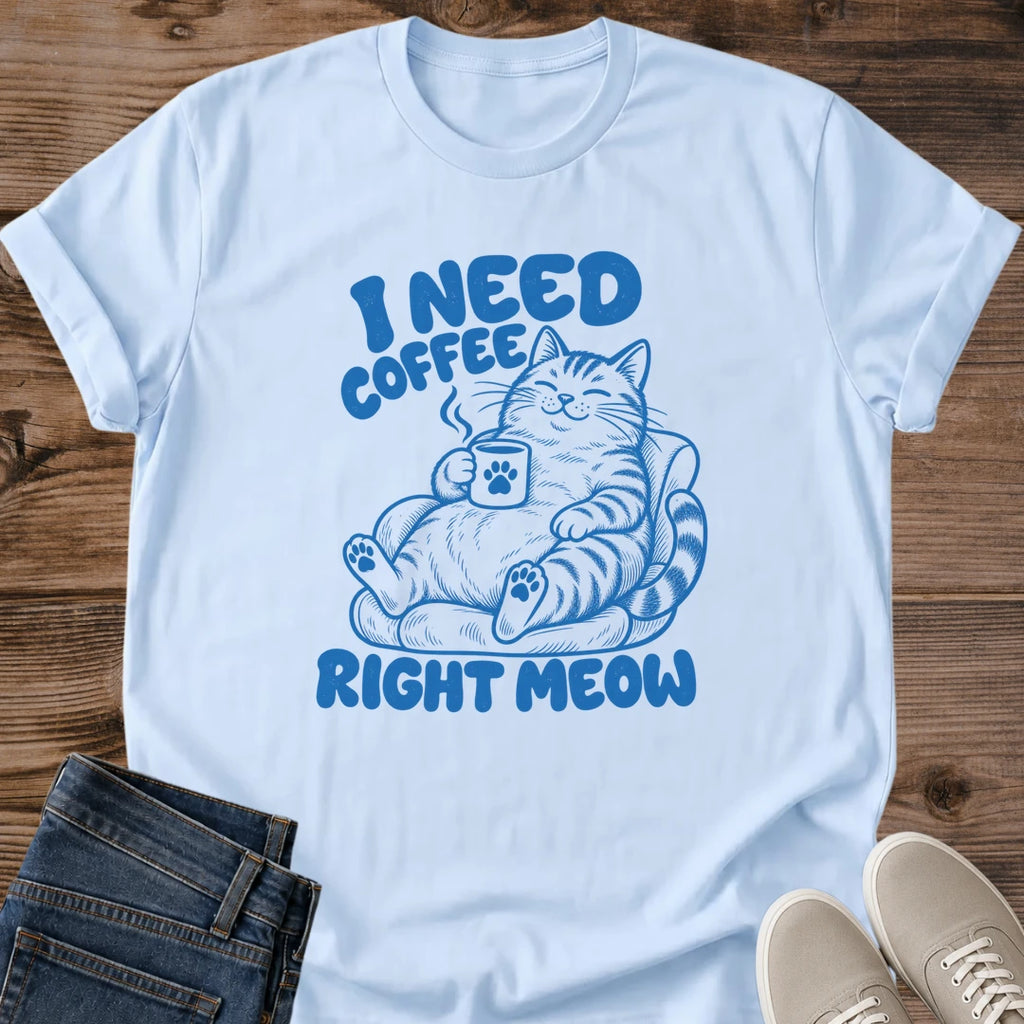Light blue T-shirt flat lay with blue lettering reading I Need Coffee Right Meow and a tabby lounging in an armchair holding a steaming paw-print mug.