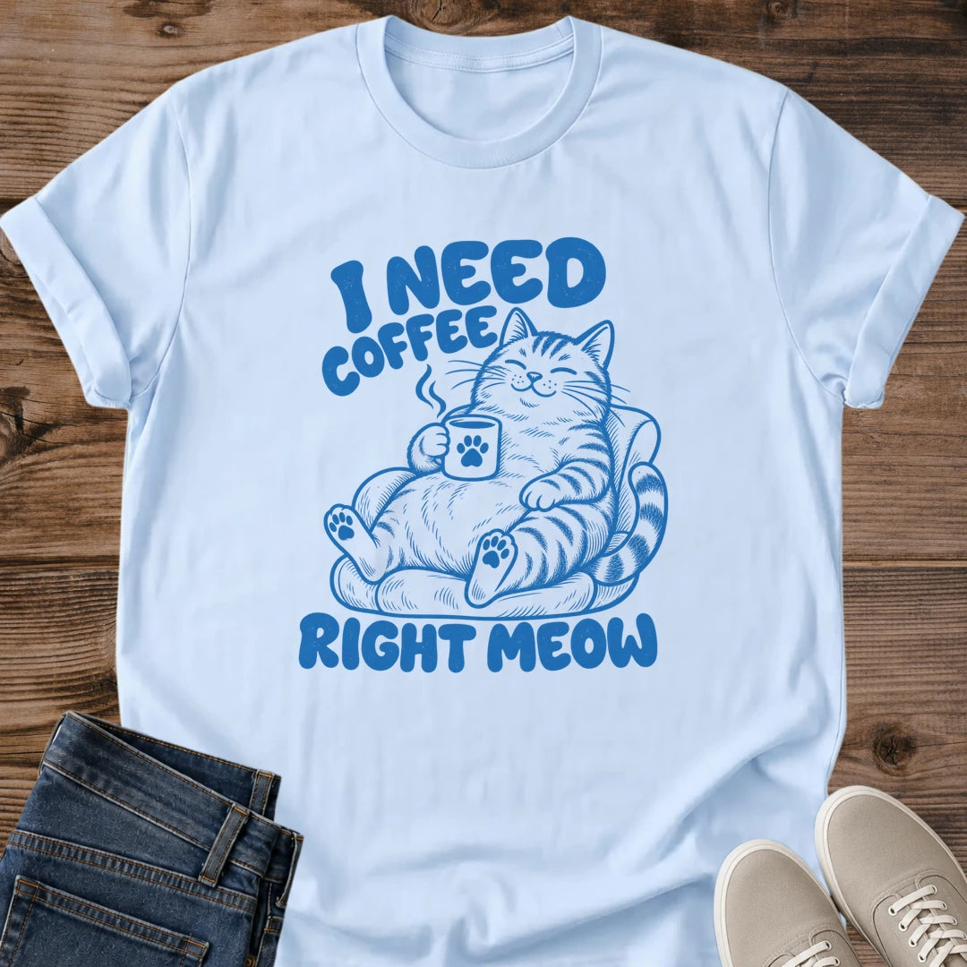 Light blue T-shirt flat lay with blue lettering reading I Need Coffee Right Meow and a tabby lounging in an armchair holding a steaming paw-print mug.