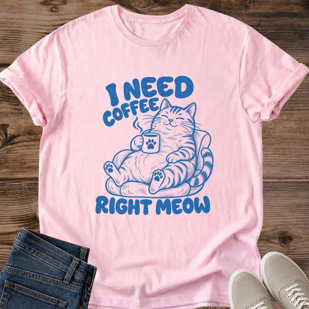 Light pink T-shirt flat lay with blue lettering reading I Need Coffee Right Meow and a tabby lounging in an armchair holding a steaming paw-print mug.