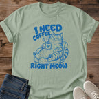 Sage green T-shirt flat lay with blue lettering reading I Need Coffee Right Meow and a tabby lounging in an armchair holding a steaming paw-print mug.