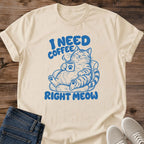 Sand T-shirt flat lay with blue lettering reading I Need Coffee Right Meow and a tabby lounging in an armchair holding a steaming paw-print mug.