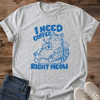 Sport grey T-shirt flat lay with blue lettering reading I Need Coffee Right Meow and a tabby lounging in an armchair holding a steaming paw-print mug.