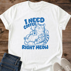 White T-shirt flat lay with blue lettering reading I Need Coffee Right Meow and a tabby lounging in an armchair holding a steaming paw-print mug.
