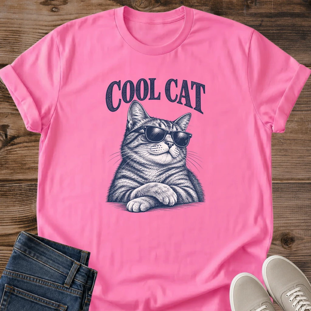Azalea bright pink Cool Cat Tabby Shades T-Shirt, a Gildan 64000 Softstyle unisex crewneck with rolled short sleeves. Navy blue illustration shows a striped tabby wearing dark sunglasses, with upright ears, long whiskers and crossed front paws beneath arched "COOL CAT" lettering. Front flat lay on rustic brown wood planks, with folded dark denim jeans at lower left and light gray lace-up sneakers at lower right.