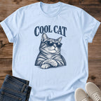 Light Blue pastel blue Cool Cat Tabby Shades T-Shirt, a Gildan 64000 Softstyle unisex crewneck with rolled short sleeves. Navy blue illustration shows a striped tabby wearing dark sunglasses, with upright ears, long whiskers and crossed front paws beneath arched "COOL CAT" lettering. Front flat lay on rustic brown wood planks, with folded dark denim jeans at lower left and light gray lace-up sneakers at lower right.