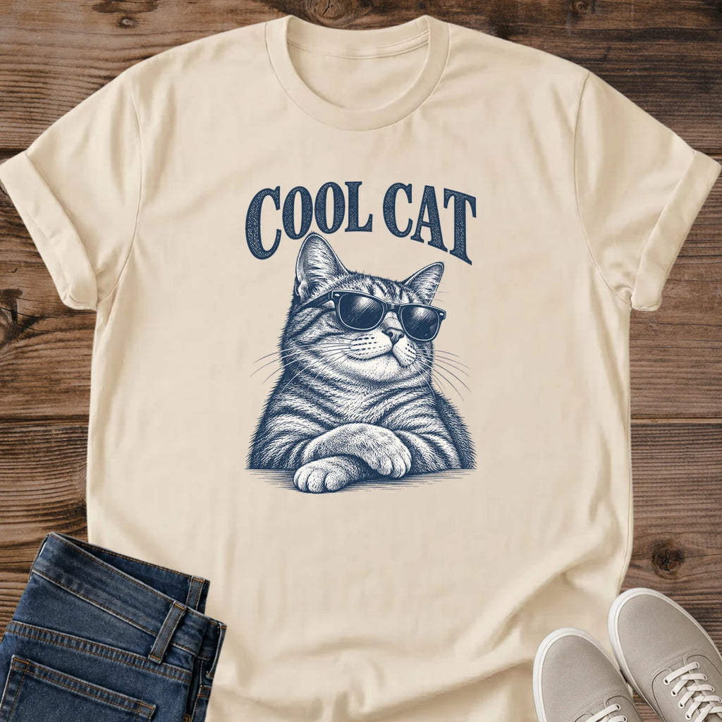 Sand beige Cool Cat Tabby Shades T-Shirt, a Gildan 64000 Softstyle unisex crewneck with rolled short sleeves. Navy blue illustration shows a striped tabby wearing dark sunglasses, with upright ears, long whiskers and crossed front paws beneath arched "COOL CAT" lettering. Front flat lay on rustic brown wood planks, with folded dark denim jeans at lower left and light gray lace-up sneakers at lower right.
