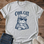 Sport Grey heather gray Cool Cat Tabby Shades T-Shirt, a Gildan 64000 Softstyle unisex crewneck with rolled short sleeves. Navy blue illustration shows a striped tabby wearing dark sunglasses, with upright ears, long whiskers and crossed front paws beneath arched "COOL CAT" lettering. Front flat lay on rustic brown wood planks, with folded dark denim jeans at lower left and light gray lace-up sneakers at lower right.