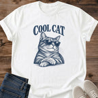 White Cool Cat Tabby Shades T-Shirt, a Gildan 64000 Softstyle unisex crewneck with rolled short sleeves. Navy blue illustration shows a striped tabby wearing dark sunglasses, with upright ears, long whiskers and crossed front paws beneath arched "COOL CAT" lettering. Front flat lay on rustic brown wood planks, with folded dark denim jeans at lower left and light gray lace-up sneakers at lower right.