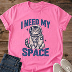 Azalea pink Gildan 64000 unisex Cosmic Kitten cat T-shirt laid flat on rustic wood with rolled sleeves, folded dark blue jeans and light gray sneakers. The front graphic reads "I NEED MY SPACE" in large distressed blue letters around a seated tabby kitten in a cream and navy astronaut suit. Its round helmet reveals its face; chest controls, a ringed-planet sleeve patch, striped tail and two small starbursts complete the illustration.