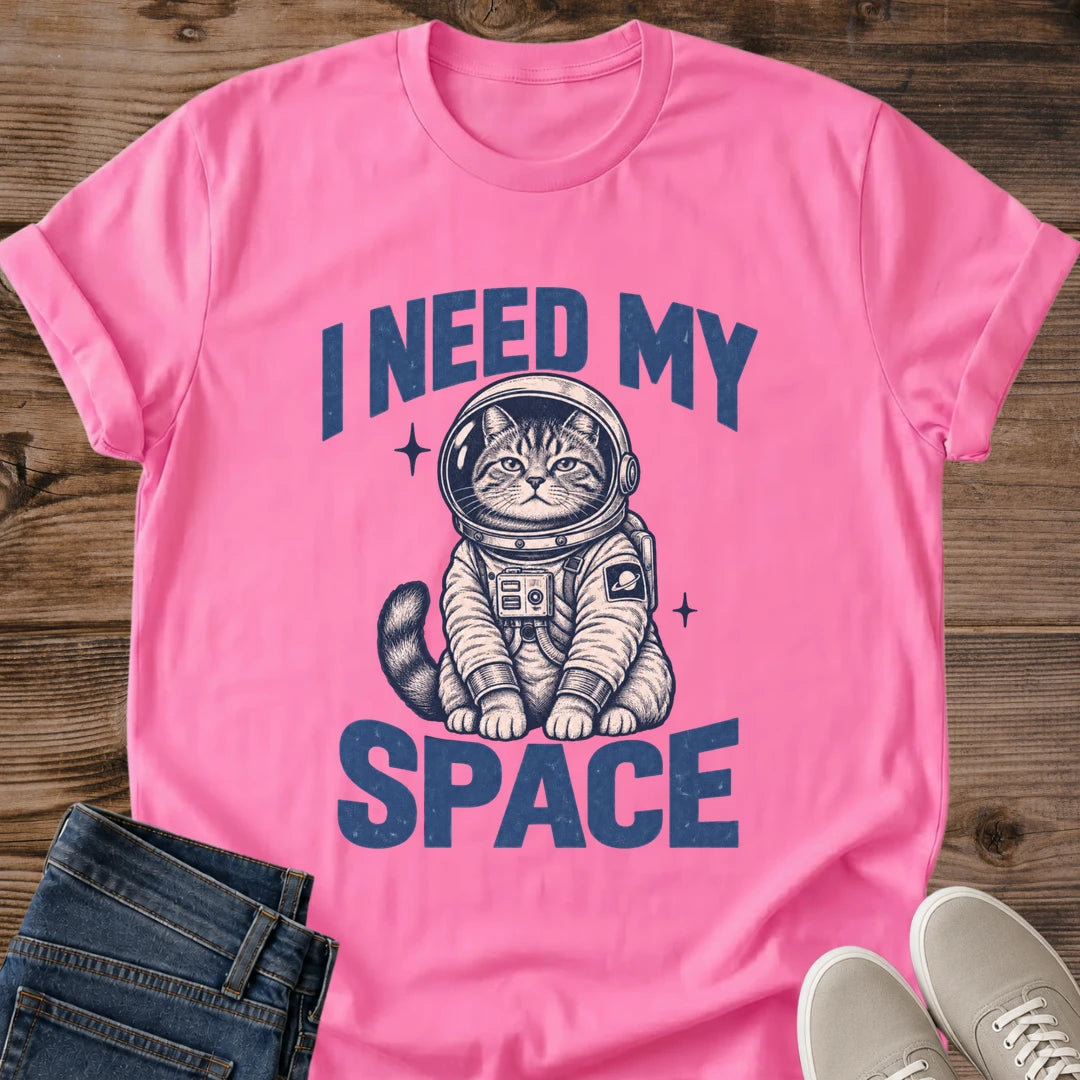 Azalea pink Gildan 64000 unisex Cosmic Kitten cat T-shirt laid flat on rustic wood with rolled sleeves, folded dark blue jeans and light gray sneakers. The front graphic reads "I NEED MY SPACE" in large distressed blue letters around a seated tabby kitten in a cream and navy astronaut suit. Its round helmet reveals its face; chest controls, a ringed-planet sleeve patch, striped tail and two small starbursts complete the illustration.