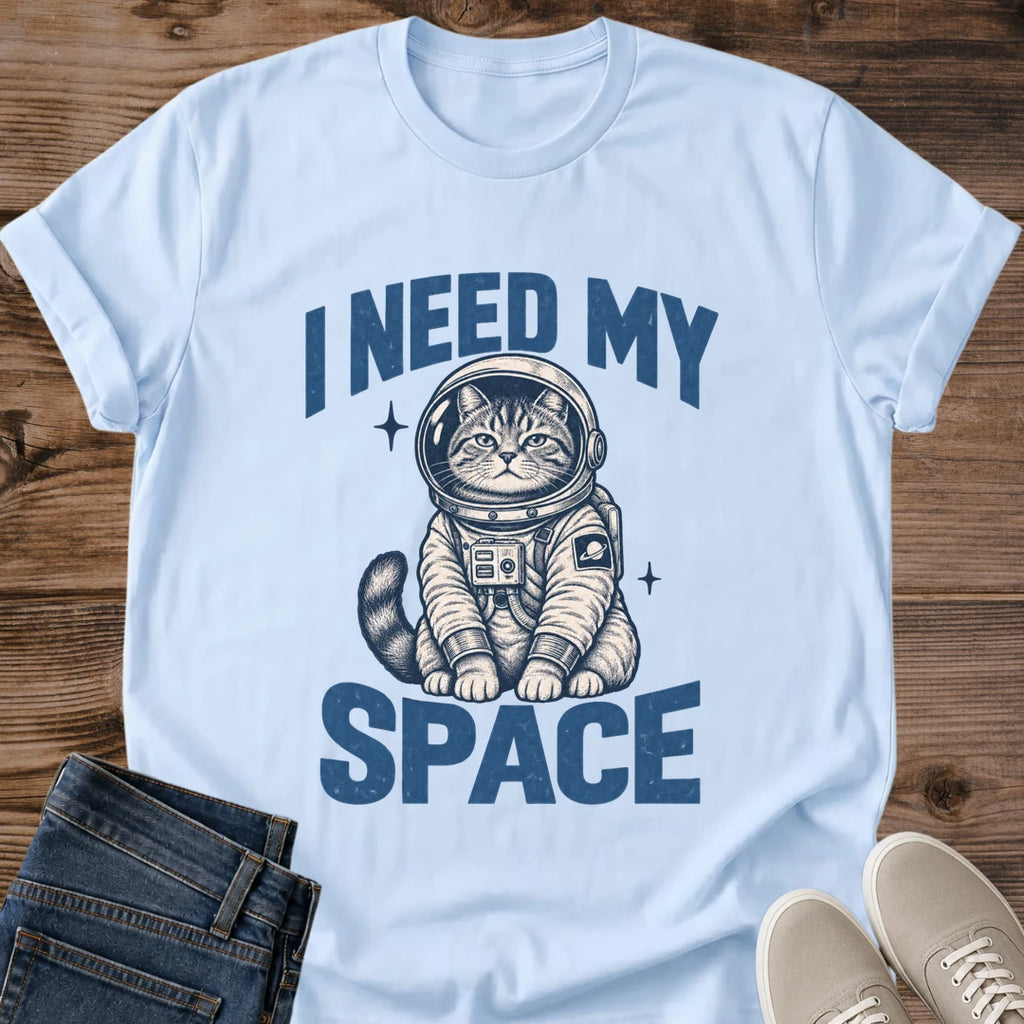 Light Blue Gildan 64000 unisex Cosmic Kitten cat T-shirt in a front-view flat lay on rustic wood beside folded blue jeans and light gray sneakers. Blue "I NEED MY SPACE" lettering frames a seated tabby kitten wearing a cream spacesuit with navy shading, a round helmet, chest controls and a ringed-planet patch. Two small starbursts flank the kitten, whose striped tail curls to one side. The pastel blue crewneck has rolled short sleeves.