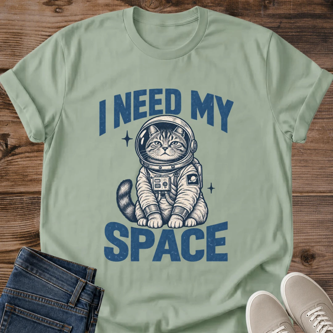 Sage green Gildan 64000 unisex Cosmic Kitten cat T-shirt shown from the front on rustic wood with folded dark jeans and light gray sneakers. The muted green crewneck has rolled sleeves and blue "I NEED MY SPACE" lettering around a seated tabby wearing a round astronaut helmet and cream spacesuit with navy detailing. Chest controls, a ringed-planet sleeve patch, a striped tail and two small starbursts are visible in the centered print.
