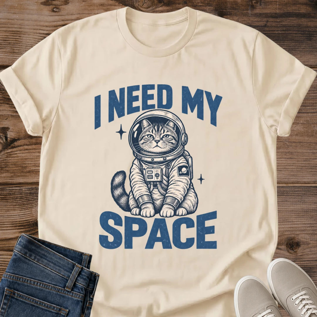Sand beige Gildan 64000 unisex Cosmic Kitten astronaut cat T-shirt shown from the front on wooden planks with a crew neck and rolled sleeves. A seated tabby in a round helmet and detailed cream spacesuit sits between blue "I NEED MY" and "SPACE" lettering. Navy linework defines its whiskers, striped tail, chest control panel and ringed-planet sleeve patch, with two starbursts nearby. Folded dark jeans and light gray lace-up sneakers sit at the bottom.