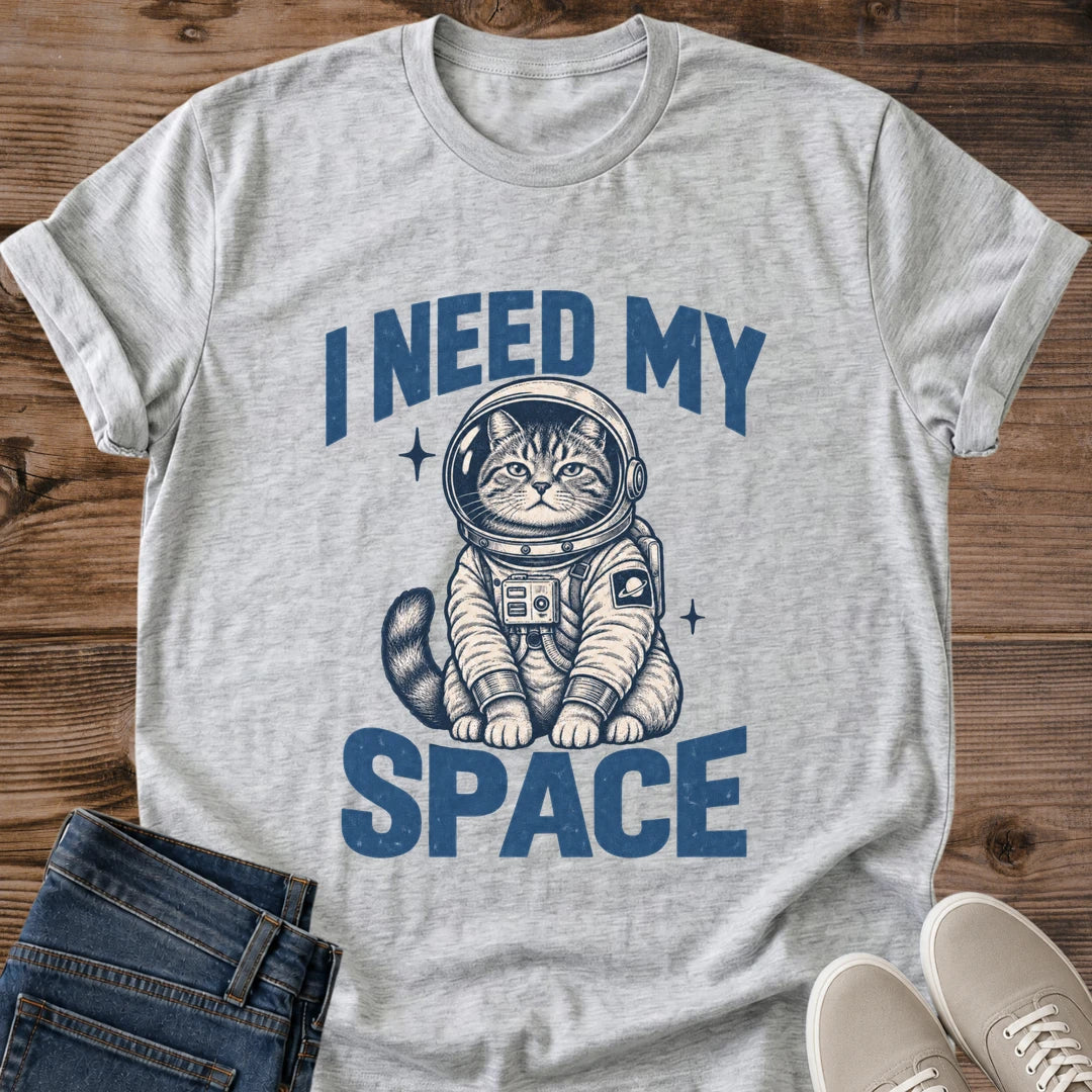 Sport Grey Gildan 64000 unisex Cosmic Kitten astronaut cat T-shirt laid flat on rustic wooden planks with rolled short sleeves. Distressed blue block letters read "I NEED MY" above a seated tabby kitten in a round space helmet and "SPACE" below. The cream and navy illustration includes a chest control panel, a ringed-planet sleeve patch, a striped tail and two starbursts. Folded dark blue jeans and light gray lace-up sneakers frame the heather gray tee.