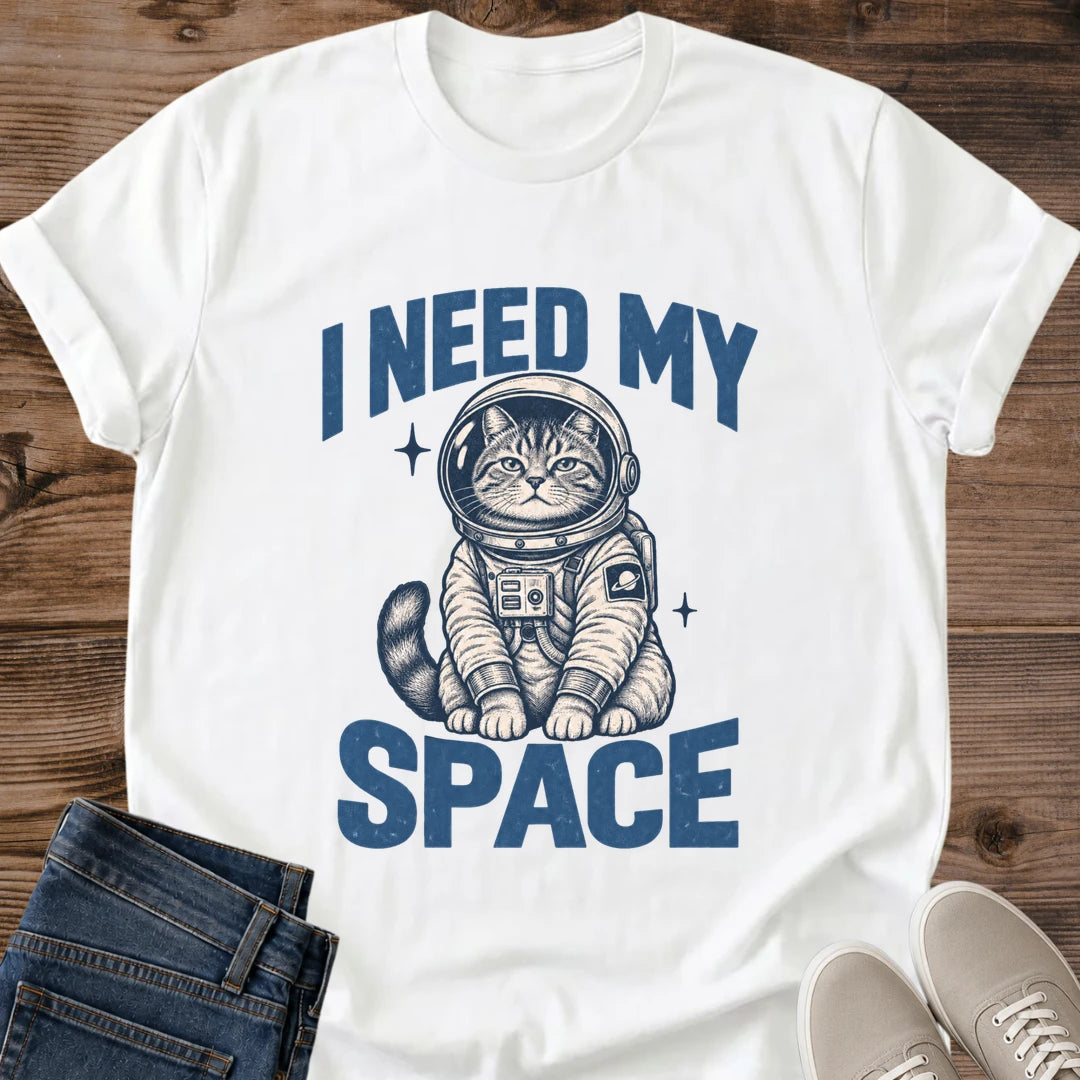 White Gildan 64000 unisex Cosmic Kitten astronaut cat T-shirt in a flat lay on wooden planks, styled with folded blue jeans and light gray lace-up sneakers. Blue block lettering reads "I NEED MY" above a seated tabby kitten and "SPACE" below. The cream and navy spacesuit has a round helmet, chest control panel and ringed-planet sleeve patch; two starbursts and a curved striped tail surround the figure. The crewneck tee has rolled short sleeves.