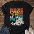 Black Gildan 64000 unisex Extra Fries cat T-shirt shown from the front on wooden planks, styled with folded blue jeans and light gray sneakers. Orange, yellow, cream and teal block lettering reads "EXERCISE? I THOUGHT YOU SAID EXTRA FRIES," with horizontal retro stripes and a distressed texture. A reclining cream tabby with closed eyes, visible paw pads and a curved striped tail sits beneath the slogan. The crewneck tee has rolled sleeves.