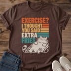 Dark Chocolate brown Gildan 64000 unisex funny cat T-shirt in a flat lay on rustic wood beside blue jeans and light gray lace-up sneakers. The rolled-sleeve crewneck features "EXERCISE? I THOUGHT YOU SAID EXTRA FRIES" in distressed orange, yellow, cream and teal block letters with matching horizontal stripes. Beneath the text, a cream striped tabby reclines with closed eyes, front paws on its belly and hind paws extended toward the viewer.
