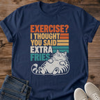 Navy blue Gildan 64000 unisex funny cat T-shirt laid flat on rustic wood with rolled short sleeves, folded dark blue jeans and light gray lace-up sneakers. The distressed retro print reads "EXERCISE? I THOUGHT YOU SAID EXTRA FRIES" in stacked orange, yellow, cream and teal lettering beside matching stripes. A cream-colored striped tabby lounges below with closed eyes, paws resting on its round belly and hind paw pads facing forward.