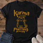 Black Feline Philosophy Cat T-Shirt, a Gildan 64000 Softstyle unisex crewneck with rolled short sleeves. Warm gold line art shows a seated smiling cat with closed eyes, round cheeks, whiskers, stripes and a curled tail, surrounded by playful lettering reading "Karma is a cat purring on my lap." Front flat lay on rustic brown wood planks, with folded dark denim jeans at lower left and light gray lace-up sneakers at lower right.
