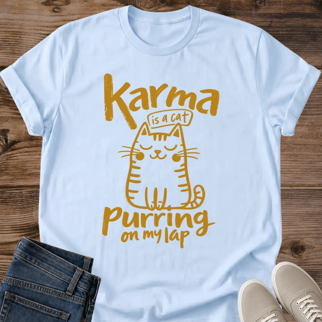 Light Blue pastel blue Feline Philosophy Cat T-Shirt, a Gildan 64000 Softstyle unisex crewneck with rolled short sleeves. Warm gold line art shows a seated smiling cat with closed eyes, round cheeks, whiskers, stripes and a curled tail, surrounded by playful lettering reading "Karma is a cat purring on my lap." Front flat lay on rustic brown wood planks, with folded dark denim jeans at lower left and light gray lace-up sneakers at lower right.