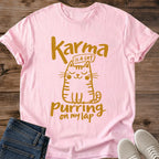 Light Pink pale pink Feline Philosophy Cat T-Shirt, a Gildan 64000 Softstyle unisex crewneck with rolled short sleeves. Warm gold line art shows a seated smiling cat with closed eyes, round cheeks, whiskers, stripes and a curled tail, surrounded by playful lettering reading "Karma is a cat purring on my lap." Front flat lay on rustic brown wood planks, with folded dark denim jeans at lower left and light gray lace-up sneakers at lower right.
