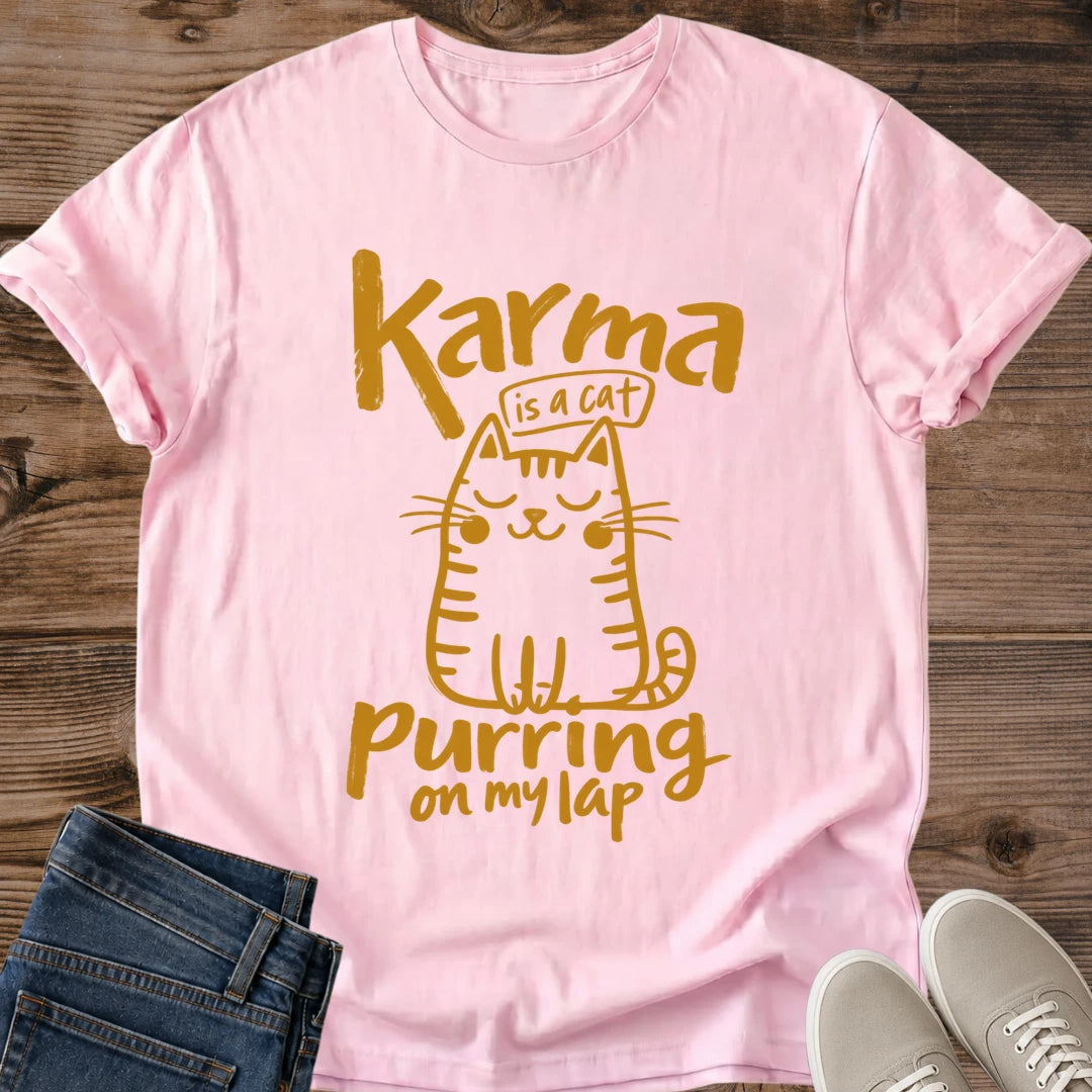 Light Pink pale pink Feline Philosophy Cat T-Shirt, a Gildan 64000 Softstyle unisex crewneck with rolled short sleeves. Warm gold line art shows a seated smiling cat with closed eyes, round cheeks, whiskers, stripes and a curled tail, surrounded by playful lettering reading "Karma is a cat purring on my lap." Front flat lay on rustic brown wood planks, with folded dark denim jeans at lower left and light gray lace-up sneakers at lower right.