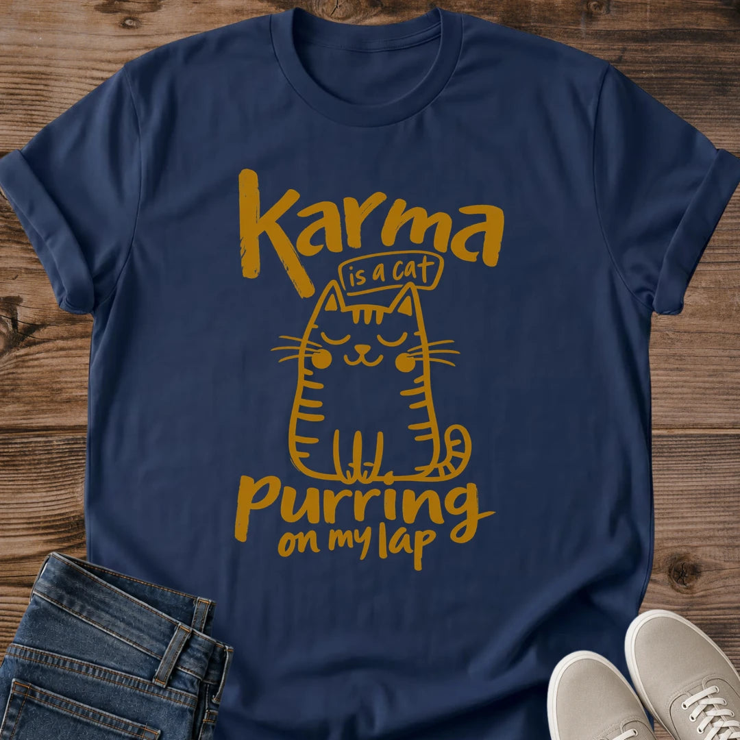 Navy dark blue Feline Philosophy Cat T-Shirt, a Gildan 64000 Softstyle unisex crewneck with rolled short sleeves. Warm gold line art shows a seated smiling cat with closed eyes, round cheeks, whiskers, stripes and a curled tail, surrounded by playful lettering reading "Karma is a cat purring on my lap." Front flat lay on rustic brown wood planks, with folded dark denim jeans at lower left and light gray lace-up sneakers at lower right.