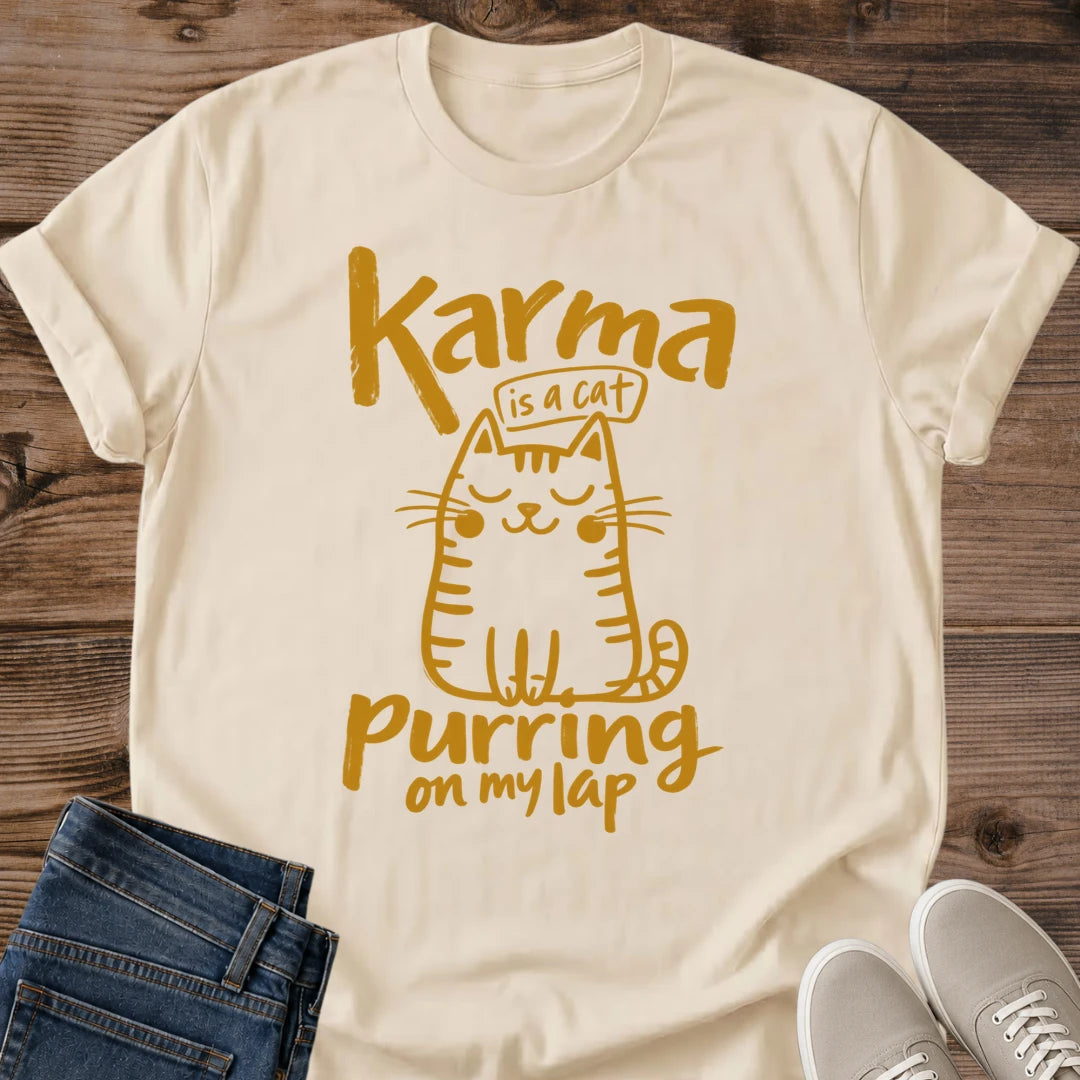 Sand beige Feline Philosophy Cat T-Shirt, a Gildan 64000 Softstyle unisex crewneck with rolled short sleeves. Warm gold line art shows a seated smiling cat with closed eyes, round cheeks, whiskers, stripes and a curled tail, surrounded by playful lettering reading "Karma is a cat purring on my lap." Front flat lay on rustic brown wood planks, with folded dark denim jeans at lower left and light gray lace-up sneakers at lower right.