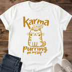 White Feline Philosophy Cat T-Shirt, a Gildan 64000 Softstyle unisex crewneck with rolled short sleeves. Warm gold line art shows a seated smiling cat with closed eyes, round cheeks, whiskers, stripes and a curled tail, surrounded by playful lettering reading "Karma is a cat purring on my lap." Front flat lay on rustic brown wood planks, with folded dark denim jeans at lower left and light gray lace-up sneakers at lower right.
