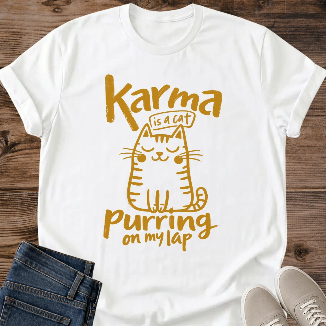 White Feline Philosophy Cat T-Shirt, a Gildan 64000 Softstyle unisex crewneck with rolled short sleeves. Warm gold line art shows a seated smiling cat with closed eyes, round cheeks, whiskers, stripes and a curled tail, surrounded by playful lettering reading "Karma is a cat purring on my lap." Front flat lay on rustic brown wood planks, with folded dark denim jeans at lower left and light gray lace-up sneakers at lower right.