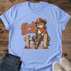 Carolina Blue Gildan 64000 unisex Howdy Darlin cat T-shirt in a flat lay on rustic wood with rolled short sleeves and a round crew neck. The centered western print shows a striped tabby seated on a wooden fence wearing a brown cowboy hat, rust-red bandana, blue jeans, fringed chaps and boots. Rust-orange "Howdy Darlin" lettering with cream edging appears beside the cat. Folded dark blue jeans and light gray lace-up sneakers frame the shirt.