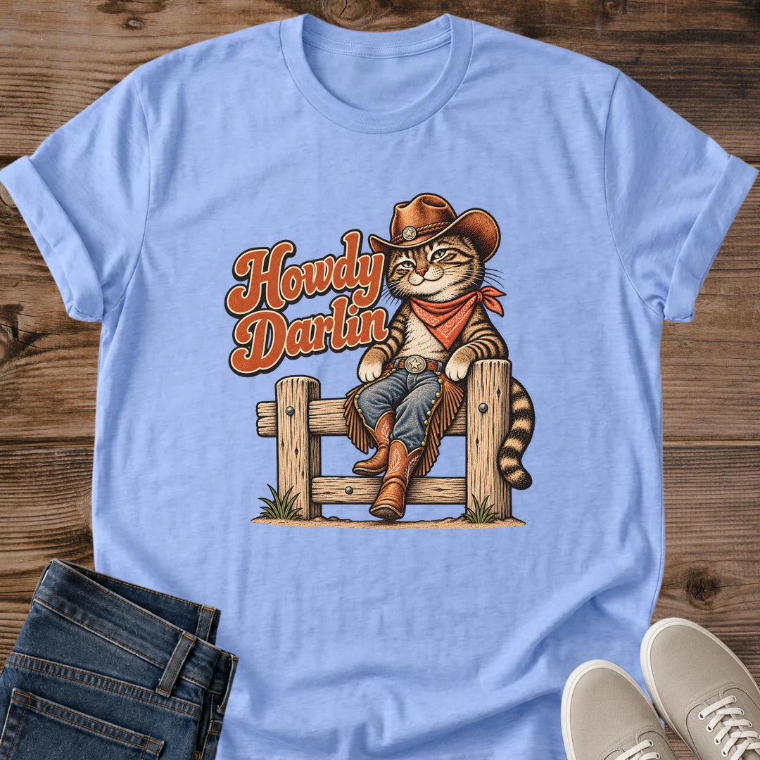 Carolina Blue Gildan 64000 unisex Howdy Darlin cat T-shirt in a flat lay on rustic wood with rolled short sleeves and a round crew neck. The centered western print shows a striped tabby seated on a wooden fence wearing a brown cowboy hat, rust-red bandana, blue jeans, fringed chaps and boots. Rust-orange "Howdy Darlin" lettering with cream edging appears beside the cat. Folded dark blue jeans and light gray lace-up sneakers frame the shirt.