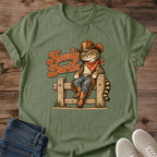Military Green olive Gildan 64000 unisex Howdy Darlin cowboy cat T-shirt shown from the front on wooden planks, styled with folded blue jeans and light gray lace-up sneakers. The rolled-sleeve crewneck features a tabby resting on a wooden fence in a brown hat, rust-red bandana, blue jeans, fringed chaps and cowboy boots. Rust-orange "Howdy Darlin" script, a striped tail and small grass tufts complete the vintage western illustration.