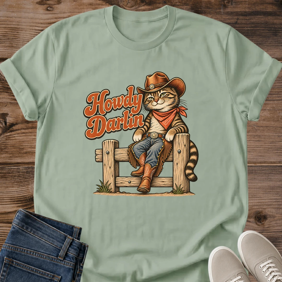 Sage green Gildan 64000 unisex Howdy Darlin cat T-shirt shown from the front on rustic wood, with a crew neck and rolled sleeves. A tabby cowboy lounges on a wooden fence wearing a brown hat, rust-red neckerchief, blue jeans, fringed chaps and brown boots, with its striped tail hanging beside a post. Rust-orange "Howdy Darlin" lettering has cream and dark outlines. Folded blue jeans and light gray sneakers complete the flat lay.