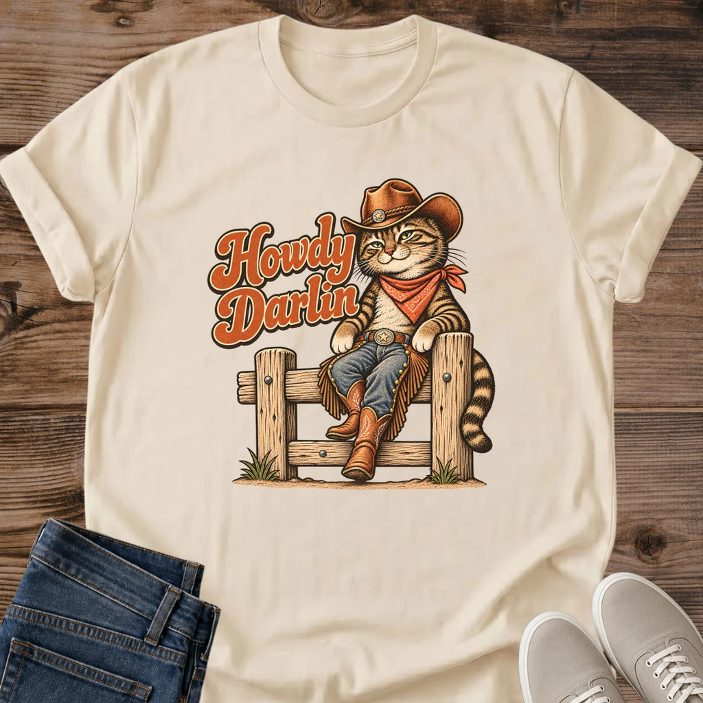 Sand beige Gildan 64000 unisex Howdy Darlin cowboy cat T-shirt laid flat on rustic wooden planks with rolled short sleeves. The vintage-style graphic shows a striped tabby sitting on a wooden fence in a brown cowboy hat, rust-red bandana, blue jeans, fringed chaps and brown boots. Rust-orange "Howdy Darlin" script with cream edging appears beside the cat. Folded dark blue jeans and light gray lace-up sneakers frame the lower corners of the crewneck tee.