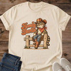 Sand beige Gildan 64000 unisex Howdy Darlin cowboy cat T-shirt laid flat on rustic wooden planks with rolled short sleeves. The vintage-style graphic shows a striped tabby sitting on a wooden fence in a brown cowboy hat, rust-red bandana, blue jeans, fringed chaps and brown boots. Rust-orange "Howdy Darlin" script with cream edging appears beside the cat. Folded dark blue jeans and light gray lace-up sneakers frame the lower corners of the crewneck tee.
