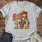 Sport Grey Gildan 64000 unisex Howdy Darlin cowboy cat T-shirt in a flat lay on wooden planks beside folded blue jeans and light gray lace-up sneakers. The heather gray crewneck has rolled sleeves and a vintage illustration of a tabby perched on a fence in a brown cowboy hat, rust-red bandana, blue jeans, fringed chaps and boots. Rust-orange "Howdy Darlin" script sits to the left, with small tufts of grass at the fence posts.