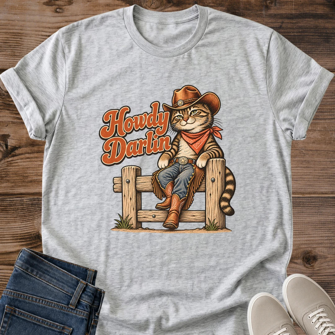 Sport Grey Gildan 64000 unisex Howdy Darlin cowboy cat T-shirt in a flat lay on wooden planks beside folded blue jeans and light gray lace-up sneakers. The heather gray crewneck has rolled sleeves and a vintage illustration of a tabby perched on a fence in a brown cowboy hat, rust-red bandana, blue jeans, fringed chaps and boots. Rust-orange "Howdy Darlin" script sits to the left, with small tufts of grass at the fence posts.
