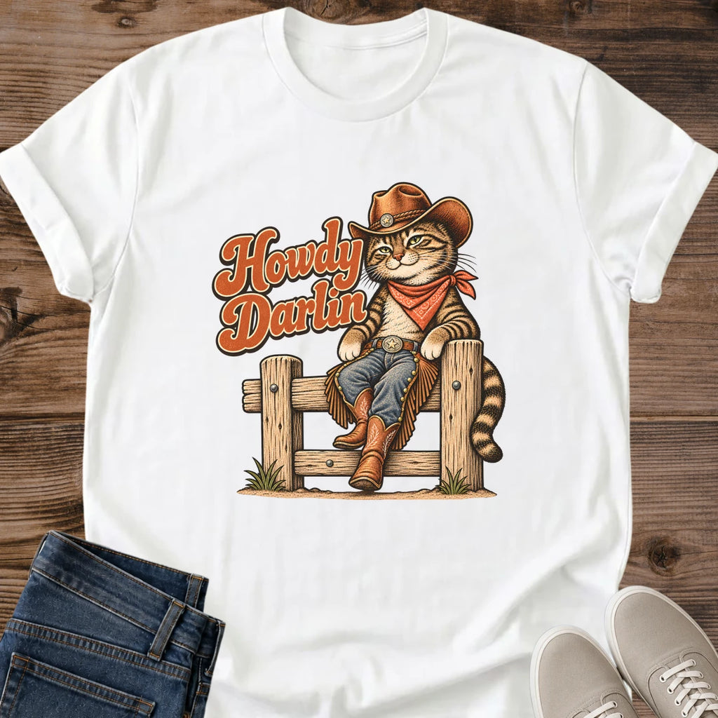 White Gildan 64000 unisex Howdy Darlin cat T-shirt laid flat on rustic wood with a crew neck and rolled short sleeves. A brown striped tabby sits on a wooden fence, dressed in a cowboy hat, rust-red bandana, blue jeans, fringed chaps and brown boots. Its tail curves beside the right fence post, and rust-orange "Howdy Darlin" script with cream edging fills the left side of the print. Folded dark jeans and light gray sneakers sit below.